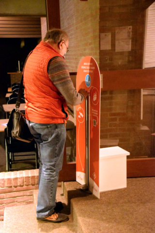 Un oeuvre, vêtu d'une veste sans manches rouge, d'une jeans, portant une besace en cuir noir, devant le présentoir à gel hydroalcoolique installé à l'entrée de l'exposition
